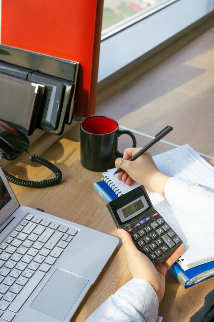 A person writing notes at a desk with a calculator, laptop, and red mug.